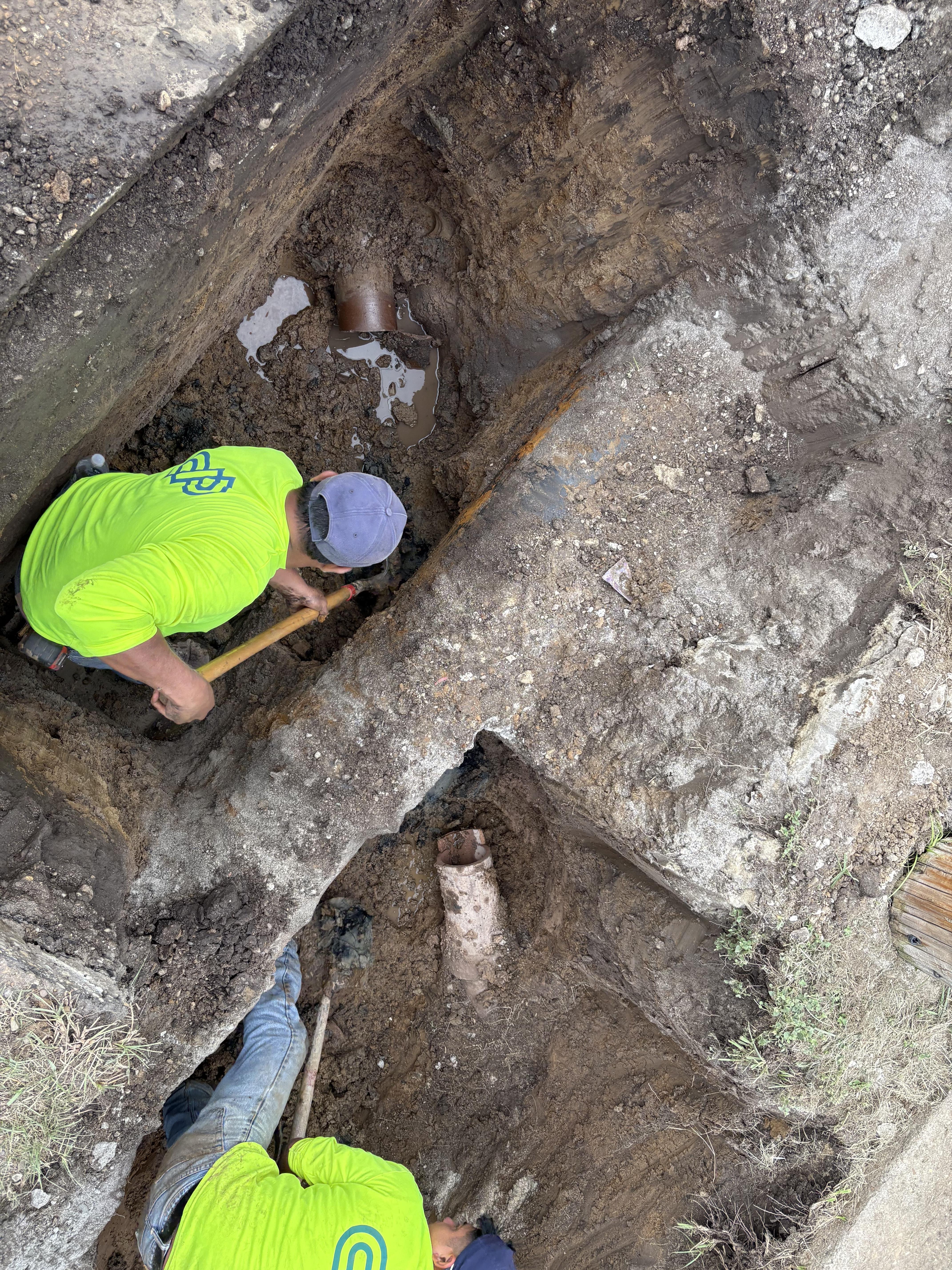 Two workers in bright yellow shirts are excavating a trench filled with dirt and mud. Visible within the trench are sections of exposed pipes partially submerged in water. The workers use tools, including shovels, to clear the area and inspect the underground infrastructure. The trench edges show layers of soil, highlighting the ongoing repair or construction activity. 