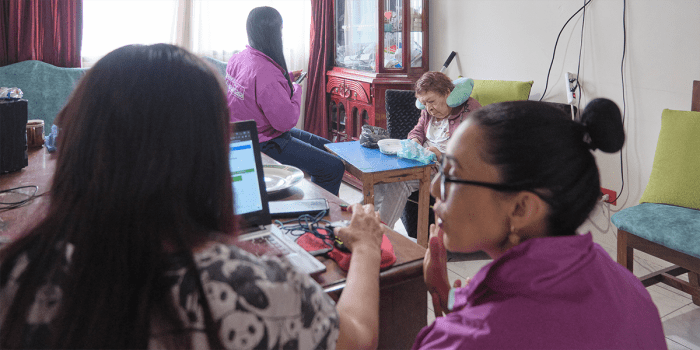 A group of three caregivers tend to an elderly patient. Two are in the foreground working on a laptop and having a conversation. The other is in the background with the patient, who has a pillow around her neck.