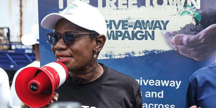 A woman wearing a white cap and sunglasses speaks into a red and white megaphone at a tree giveaway campaign event. She is dressed in a black t-shirt and has a microphone clipped to her collar. Behind her, a large banner with bold text and an image of a hand holding a small tree sapling promotes the initiative.