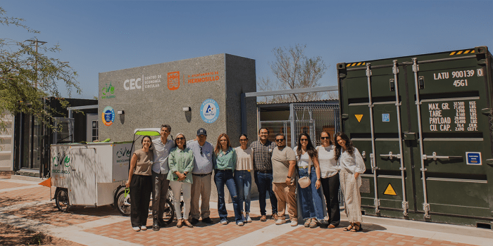 A group of people poses in front of the "Centro de Economía Circular" (Circular Economy Center) in Hermosillo, Mexico. They stand on a paved area under a clear blue sky, smiling at the camera. To the left, a white cargo tricycle labeled "Biciclando" is parked. On the right, a large green shipping container with weight and capacity markings is visible. The background features the modern, gray building of the center, adorned with various logos, including the Hermosillo municipal emblem and sustainability-rela