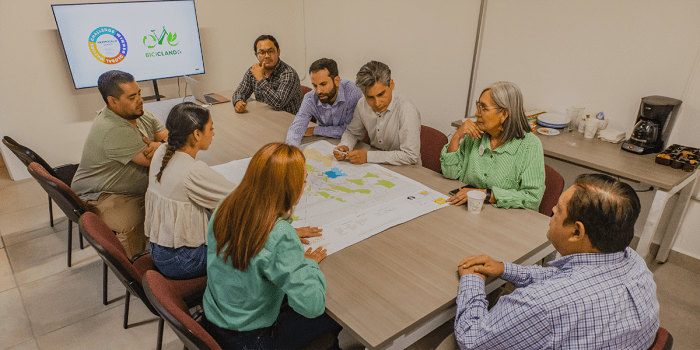 A group of eight people is gathered around a table in a meeting room, discussing a large map spread out before them.