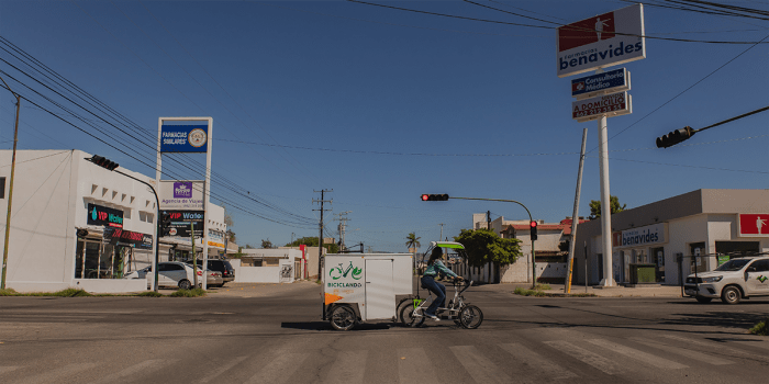 A person rides a cargo tricycle labeled "Biciclando" through an intersection in Hermosillo, Mexico, under a clear blue sky. The cyclist wears a green jacket and dark pants, pedaling while waiting for the red traffic light.