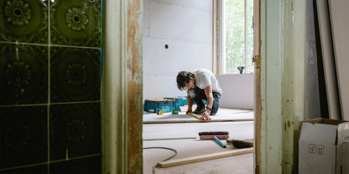 A construction worker in a white t-shirt and dark pants kneels on the floor of a partially renovated room, measuring a wooden plank. The room has exposed drywall and an open window letting in natural light. Tools, a broom, and a toolbox are scattered around. 