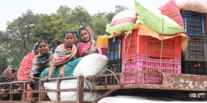 A group of women in colorful shawls and headscarves sit on the back of a truck loaded with crates of tomatoes and large sacks. Some lean on the railing, gazing ahead, while others sit closely together.