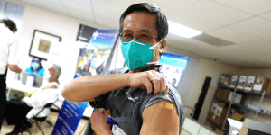 A doctor lifting up his left shoulder sleeve to show a bandage signaling that he has received a COVID-19 vaccine