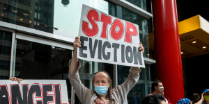 A masked man holds up a sign in front of a glass front building that reads "STOP EVICTION" and is surrounded by a group of people.