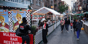 The photo features a closed off street within a city. Individuals are eating in sectioned off restaurant fronts. People are walking down the middle of the street. A person is having their photograph taken. 