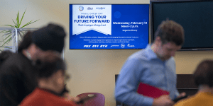 A television screen displaying the event sign for a Mobile Career Unit.  In the foreground, participants mill about, just out of focus.