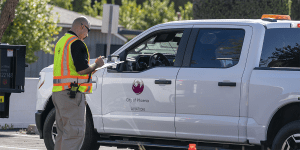 A security officer wearing a safety vest with a radio on his hip.  He is holding a clipboard and talking to the driver of a white truck with a City of Phoenix Aviation logo on the side.