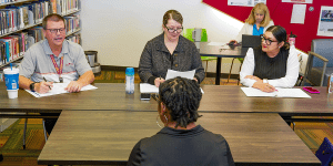 Three interviewers talk to a candidate.  Two narrow tables are between the interviewers and the candidate.