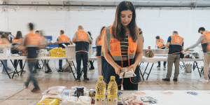 The image depicts a young woman wearing an orange safety vest in a warehouse, sorting and organizing food supplies such as bottled cooking oil, packaged grains, and canned goods. Other volunteers in similar vests are working in the background, some appearing blurred due to movement.