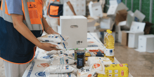 A volunteer wearing an orange and navy blue vest is packing food items into a box at a distribution center. The table is covered with staple foods such as rice, beans, pasta, cooking oil, and canned goods. In the background, there are other volunteers working and stacks of cardboard boxes.