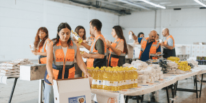 The image shows a group of volunteers wearing orange safety vests working in a warehouse setting. They are organizing and packing food supplies, including bottled cooking oil, bags of rice, and other non-perishable goods. 