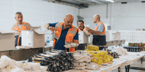 Several volunteers wearing orange vests are assembling food packages at a distribution center. They are filling cardboard boxes with essential food items such as pasta, rice, beans, and canned goods. The background shows additional volunteers and stacks of packed boxes.