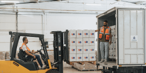 A worker operating a yellow forklift is loading a pallet of neatly stacked white boxes into a truck. Another worker, standing inside the truck, is guiding the placement of the load. Both are wearing orange safety vests.