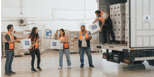 A group of workers in a warehouse is unloading white boxes from a truck, forming a human chain to pass them along efficiently. They are wearing high-visibility orange vests.