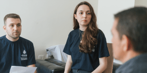 Two individuals wearing navy blue shirts with 'İstanbul Büyükşehir Belediyesi' (Istanbul Metropolitan Municipality) and 'Sosyal Hizmetler Şube Müdürlüğü' (Social Services Branch Directorate) logos sit indoors, attentively listening to a third person whose back is facing the camera. 