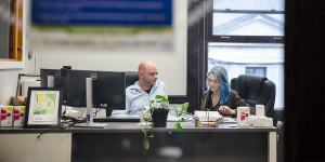 Two coworkers sit at a desk in a modern office, collaborating on paperwork. The man on the left wears a light blue shirt and leans slightly forward, looking at the woman. The woman on the right has blue hair and wears a black blazer while writing in a notebook. On the desk are two computer monitors, a potted plant, a framed print, and Narcan boxes.