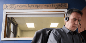 A man in the foreground is sitting at a desk.  He is wearing a headset and is slightly out of focus.  In the background, in focus, is a window with a cardboard sign above it.  The cardboard sign reads, "Do not judge people by the traps that ensnare them, but by the efforts they make to escape."  The quote is attributed to Ed Boze.