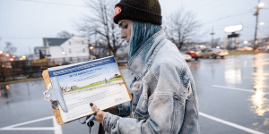 A woman with blue hair walking through the rain while wearing an acid-washed denim vest and a beanie hat.  She is holding a clipboard with a promotional document on it whose header reads, "Lift the burden of daily dosing."