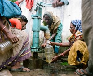 A group of people wearing head scarves and surrounding a water spigot. One of them is holding a plastic bottle underneat the tap.