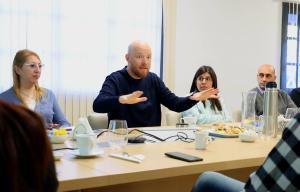 Un hombre calvo con barba roja, vestido con un suéter azul marino, gesticula con ambas manos mientras habla durante una reunión alrededor de una mesa de conferencias de madera. Cuatro colegas lo escuchan atentamente, entre ellos una mujer con lentes y cabello rubio a la izquierda, una mujer con lentes y cabello largo y oscuro con una blusa celeste, y un hombre calvo con vestimenta ejecutiva informal a la derecha.