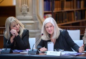 Duas mulheres de blazer preto estão sentadas em uma mesa de conferência com microfones durante uma reunião formal. A mulher à direita, com cabelo claro e comprido, gesticula enquanto fala; diante dela há uma placa com o nome “Mayor Dawn Arnold” e um copo de água. A mulher à esquerda, loira, apoia o queixo nas mãos enquanto escuta atentamente. Ao fundo, estantes de livros e detalhes arquitetônicos ornamentados sugerem uma histórica câmara governamental.