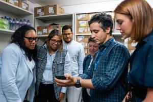 Seis profissionais se reúnem em um depósito com prateleiras de caixas de papelão e garrafas de água, concentrados na tela de um smartphone segurado por um homem de camisa xadrez azul. O grupo diverso inclui duas mulheres de óculos à esquerda, vestindo trajes profissionais claros, um homem de óculos e camisa branca ao centro, e dois outros membros da equipe à direita, todos atentos ao conteúdo exibido no celular.