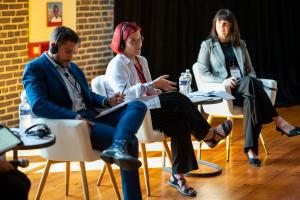 Tres panelistas se sientan en sillas blancas modernas sobre un escenario de madera durante una conversación. De izquierda a derecha: un hombre con traje azul sosteniendo documentos, una mujer con cabello rosado y lentes en vestimenta ejecutiva informal gesticulando mientras habla, y una mujer con blazer celeste sosteniendo papeles. Botellas de agua y documentos se encuentran sobre pequeñas mesas a su lado.