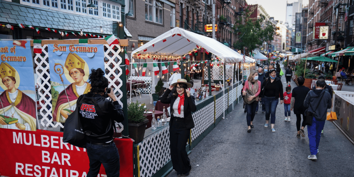The photo features a closed off street within a city. Individuals are eating in sectioned off restaurant fronts. People are walking down the middle of the street. A person is having their photograph taken.