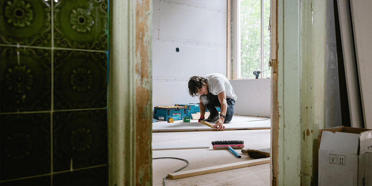 A construction worker in a white t-shirt and dark pants kneels on the floor of a partially renovated room, measuring a wooden plank. The room has exposed drywall and an open window letting in natural light. Tools, a broom, and a toolbox are scattered around. 