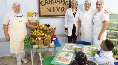 Four female school cafeteria workers smiling, standing next to a large display of fresh plant-based products on a table as part of the Sustainable School Menu program.  Two students are sitting at a school desk looking up at the women.