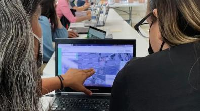 The backs of two women working at a computer together.  One woman is pointing her fingers at a map on the computer screen as a part of the Visor Urbano program.