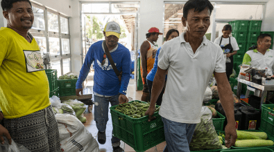 Two men carrying a carton of okra into a market.  Other customers bustle about them.