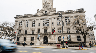 The front of a historic-looking government building.  There are three statues in front of the building, each faded to green in the elements.  Cars and people are blurred in front of the building, suggesting that the world moves extremely quickly.