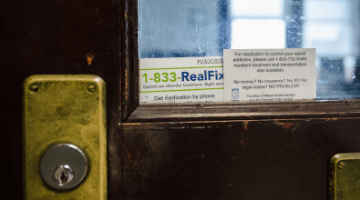 A worn wooden door with brass fittings has two business cards wedged in between the glass of the door's window and the door itself.  One card bears the phone number 1-833-RealFix.  The other gives directions on a service line for opioid addiction from the office of Mayor Andre Sayegh.