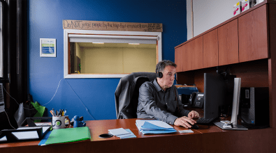 A man in the foreground is sitting at a desk.  He is wearing a headset and is slightly out of focus.  In the background, in focus, is a window with a cardboard sign above it.  The cardboard sign reads, "Do not judge people by the traps that ensnare them, but by the efforts they make to escape."  The quote is attributed to Ed Boze.