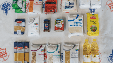 A variety of food staples are neatly arranged on a white cloth labeled "LOJİSTİK DESTEK MERKEZİ" (Logistics Support Center). The assortment includes grains like rice, lentils, and bulgur, pasta, canned tomato paste, sugar, flour, salt, tea, cooking oil, and soup mixes. The brands suggest the items are sourced from Turkish suppliers, and the packaging indicates they are intended for distribution to those in need.
