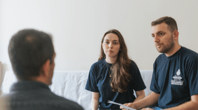 Two people wearing navy blue shirts with city logos sit across from a man, engaging in a serious discussion. The woman in the center has long brown hair and a focused expression, while the man beside her holds a piece of paper and listens attentively.