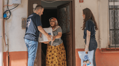 Two aid workers in navy blue shirts distribute relief supplies to a woman standing in the doorway of her home. The man hands her a cardboard box, while the woman beside him holds a plastic bag containing additional provisions. The recipient, wearing a headscarf and a long patterned skirt, accepts the aid with a serious expression, highlighting the significance of the assistance being provided.