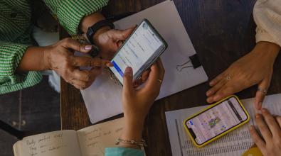 A close-up of three people working together at a wooden table, focusing on two smartphones displaying maps and route planning apps. One person wearing a green striped shirt is holding a smartphone while another person points at the screen. A second smartphone in a yellow case rests on a printed document with a list of locations. A notebook with handwritten notes and a clipboard with papers are also on the table, indicating a collaborative planning or logistics discussion.