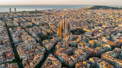 Aerial view of Barcelona, Spain, featuring the Sagrada Família basilica surrounded by dense city blocks and the Mediterranean Sea in the background.