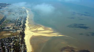 Aerial view of Beira, Mozambique, showing a long stretch of sandy coastline with tidal patterns and buildings and homes lining the shore.