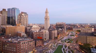 Aerial view of Boston, Massachusetts with green space, a lit street, and city buildings in soft, blue and pink light.